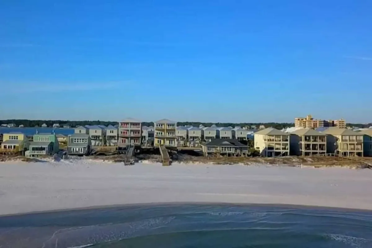 A classic Gulf Shores scene with beachfront houses directly bordering a pristine stretch of sandy beach and gentle ocean waves. The expansive sky, glistening water, and open shoreline evoke feelings of relaxation and possibility. The perspective showcases how close the homes are to the water, reinforcing the 'steps from your door' concept.