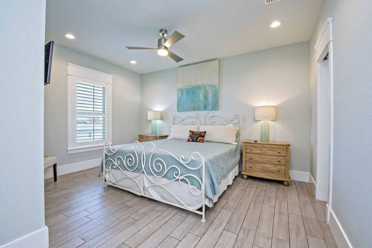 A tastefully decorated bedroom with a decorative headboard, colorful accent pillow, warm artwork above the bed, and nightstands with lamps on either side. The room exudes an inviting, cozy charm and is filled with natural light.