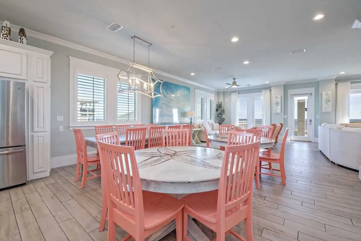 A bright dining area with a large table surrounded by colorful chairs, artwork on the wall, and abundant natural light pouring through big windows. The setting is cheerful and inviting, making it a hub for shared meals and celebrations.