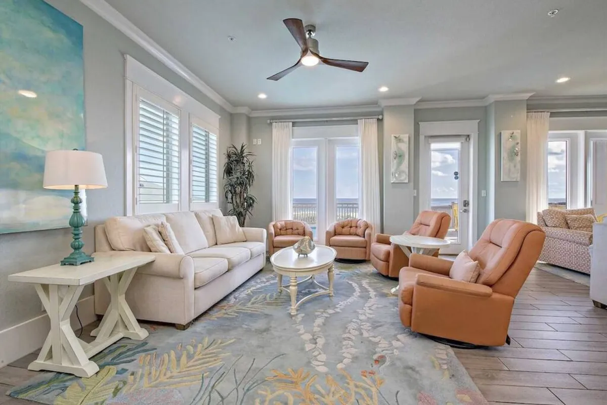 A spacious great room featuring a comfortable sofa, recliners, a coffee table, and cozy decor. Sunlight streams in through large windows, illuminating the seating area and giving the space a warm, welcoming ambiance ideal for relaxing or socializing.