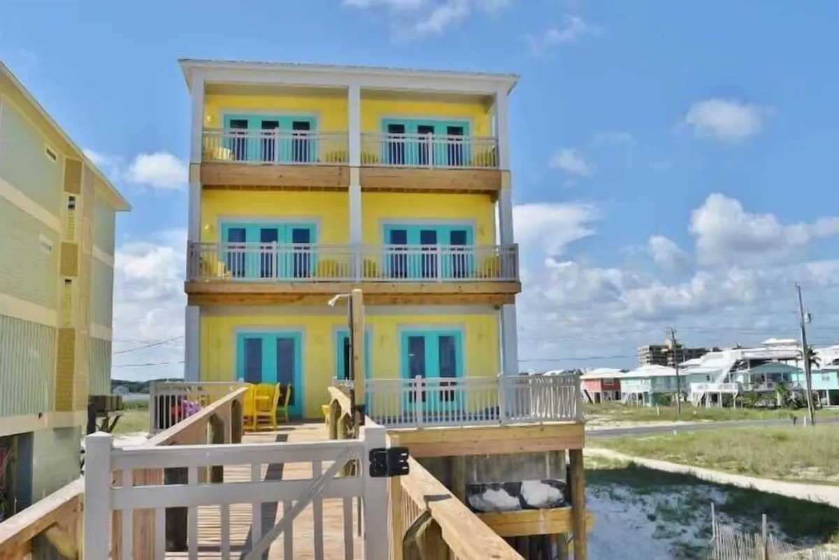 The image showcases a vibrant, yellow three-story beach house with striking blue doors and windows, multiple balconies with white railings overlooking the Gulf, and a private wooden walkway leading from the house directly to the beach. The ground-level porch features inviting outdoor seating, setting the scene for relaxation and gatherings under the sun.