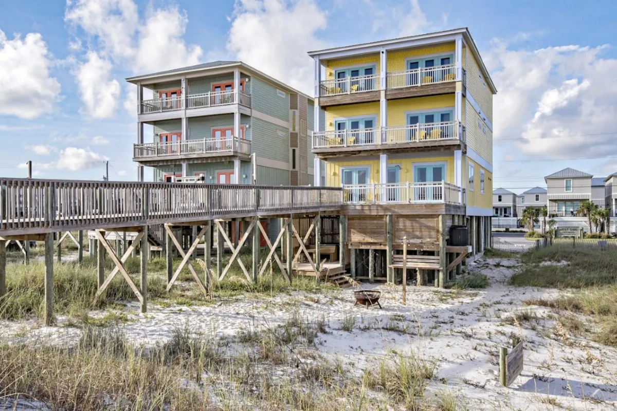 The image shows a cheerful yellow beachfront house with multiple balconies, a wooden walkway leading directly to the sandy beach, and adjacent pastel-colored beach houses. The setting is bright and inviting, with sandy dunes and sparse vegetation creating a classic coastal ambiance. This exterior view captures the proximity to the beach and the welcoming, vibrant energy of the property.