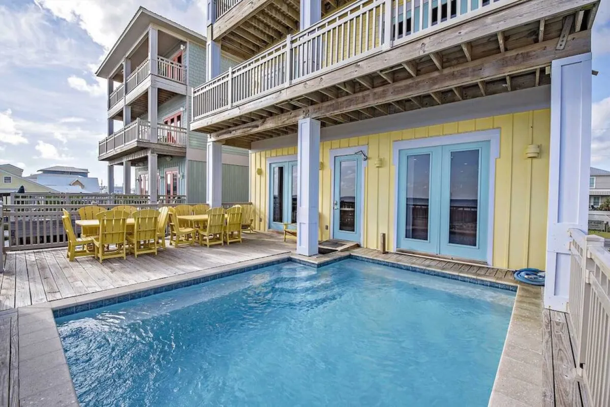 An exterior view of the bright yellow beachfront house, with balconies overlooking a private heated pool, an outdoor dining area with yellow chairs and table, and the wooden deck leading directly to the sandy beach.