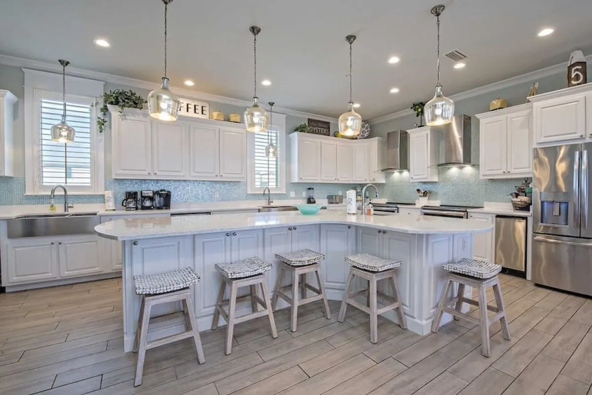 A bright, fresh kitchen scene with a large white island featuring ample seating, stainless steel refrigerator and appliances, and glass pendant lights overhead. The light blue mosaic backsplash adds a coastal touch, while the open layout allows for easy socializing and meal preparation.