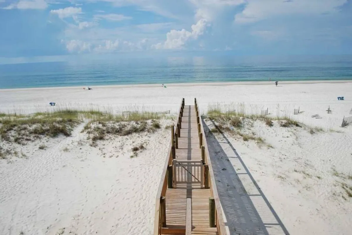 A wooden walkway stretches across the dunes, leading to a pristine white sandy beach bordered by calm ocean waters and a blue sky with scattered clouds. The image conveys a sense of peace and the promise of beachside activities.