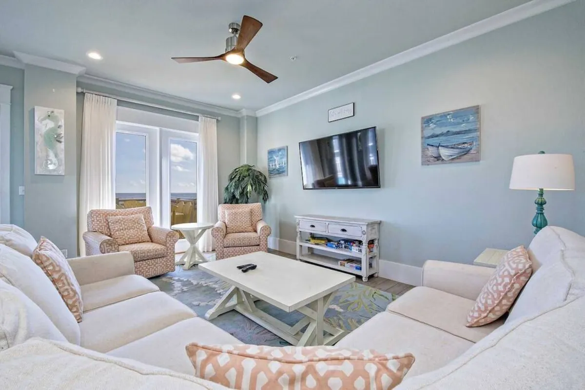 A comfortable great room featuring a large sectional sofa with patterned pillows, two accent armchairs, a wall-mounted flat screen TV, and decorative coastal paintings. The layout invites relaxation, conversation, and group activities such as game nights or movie marathons.