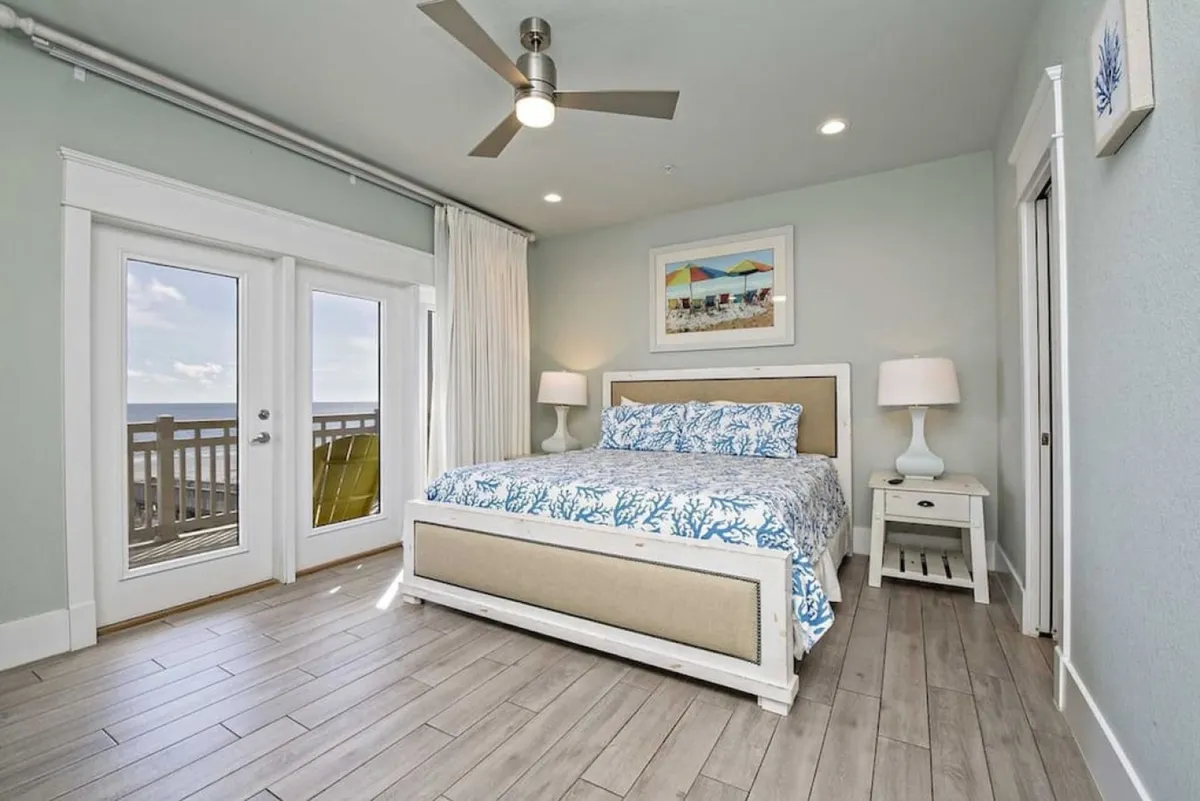 A serene king bedroom with coral-patterned bedding, flanked by matching bedside tables and lamps. French doors open directly onto a beachfront porch, allowing natural light to fill the room and offering seamless indoor-outdoor living. A ceiling fan and Smart HDTV complete the tranquil, well-appointed space.