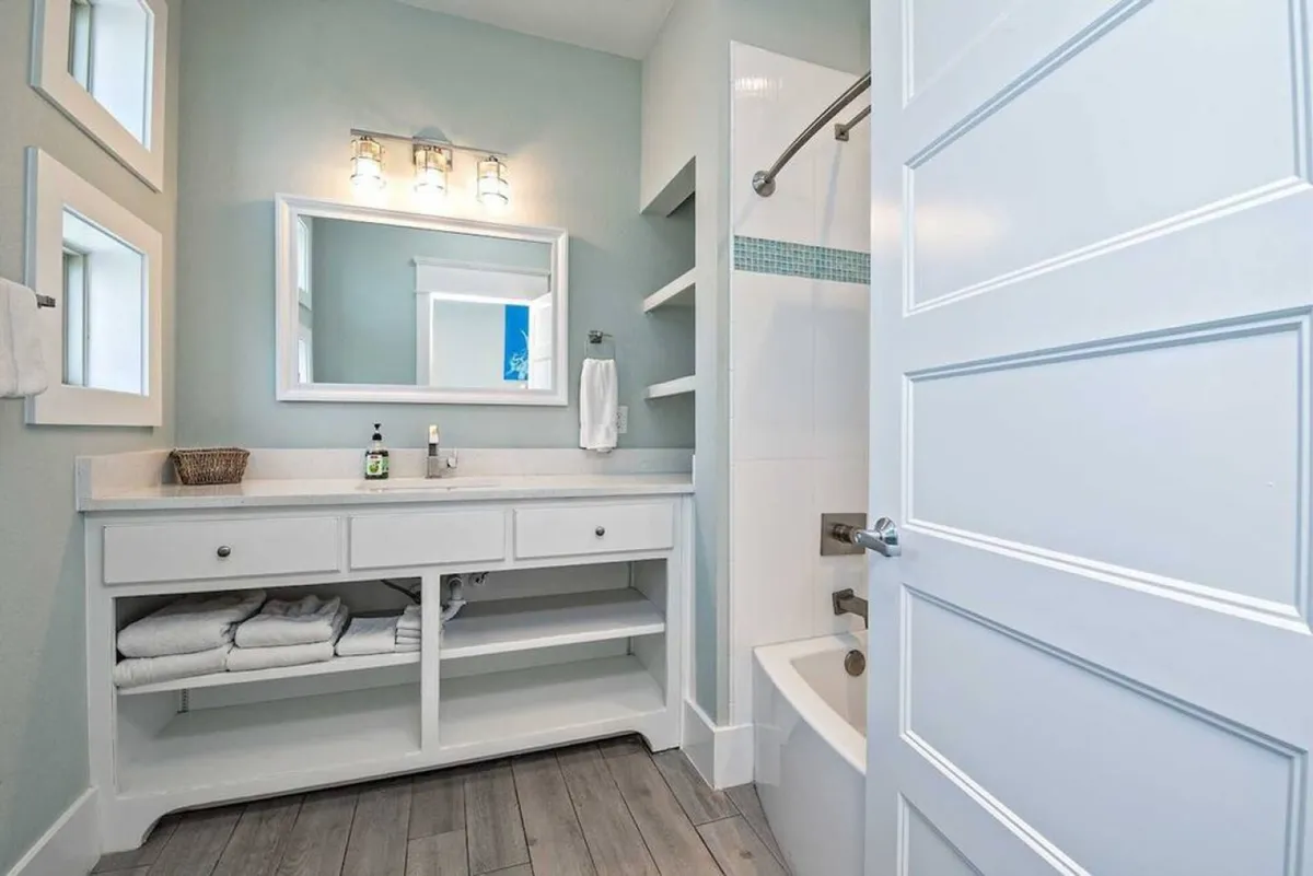 A pristine en-suite bathroom with a white vanity, open shelving for towels, a rectangular mirror with overhead lighting, and a bathtub with shower. The space is thoughtfully organized and exudes a calming, spa-like vibe.