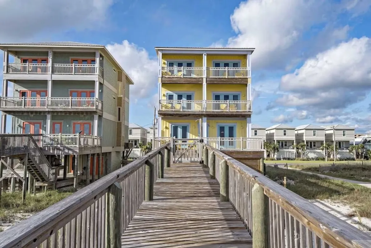 A vibrant yellow three-story beachfront house with multiple balconies overlooks the expansive beach. The wooden walkway leads directly from the house to the sugar-white sand, with clear views of the adjacent blue-gray beach house and a bright, inviting sky above.