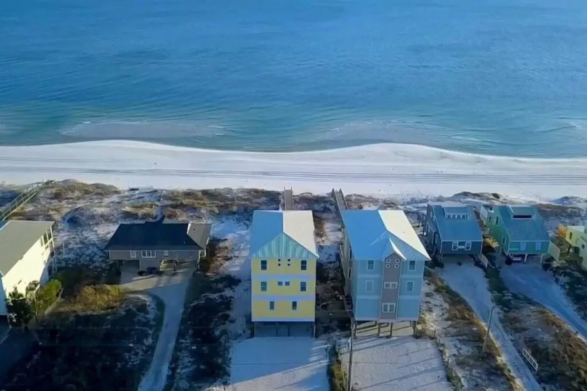 A bright yellow beachfront house with a direct dune walkover sits adjacent to colorful neighboring homes. The white sandy beach and emerald blue waters stretch out in front, with families enjoying the privacy and tranquility away from condo crowds.
