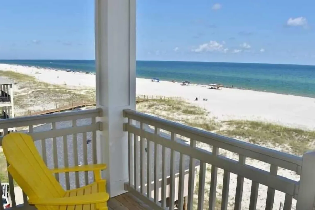 A yellow Adirondack chair sits on a private porch, framed by a white wooden railing, with a stunning view over the sandy beach and emerald-blue ocean. The scene is serene and open, inviting relaxation and sunrise or sunset watching.