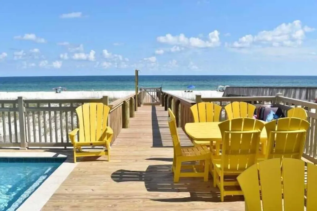 A private deck area with bright yellow Adirondack chairs and a dining table set, overlooking the ocean and white sandy beach. The pool edge is visible to the left, and the wooden walkway leads directly to the beach. The scene is sunny, tranquil, and ideal for both social gatherings and quiet moments by the water.