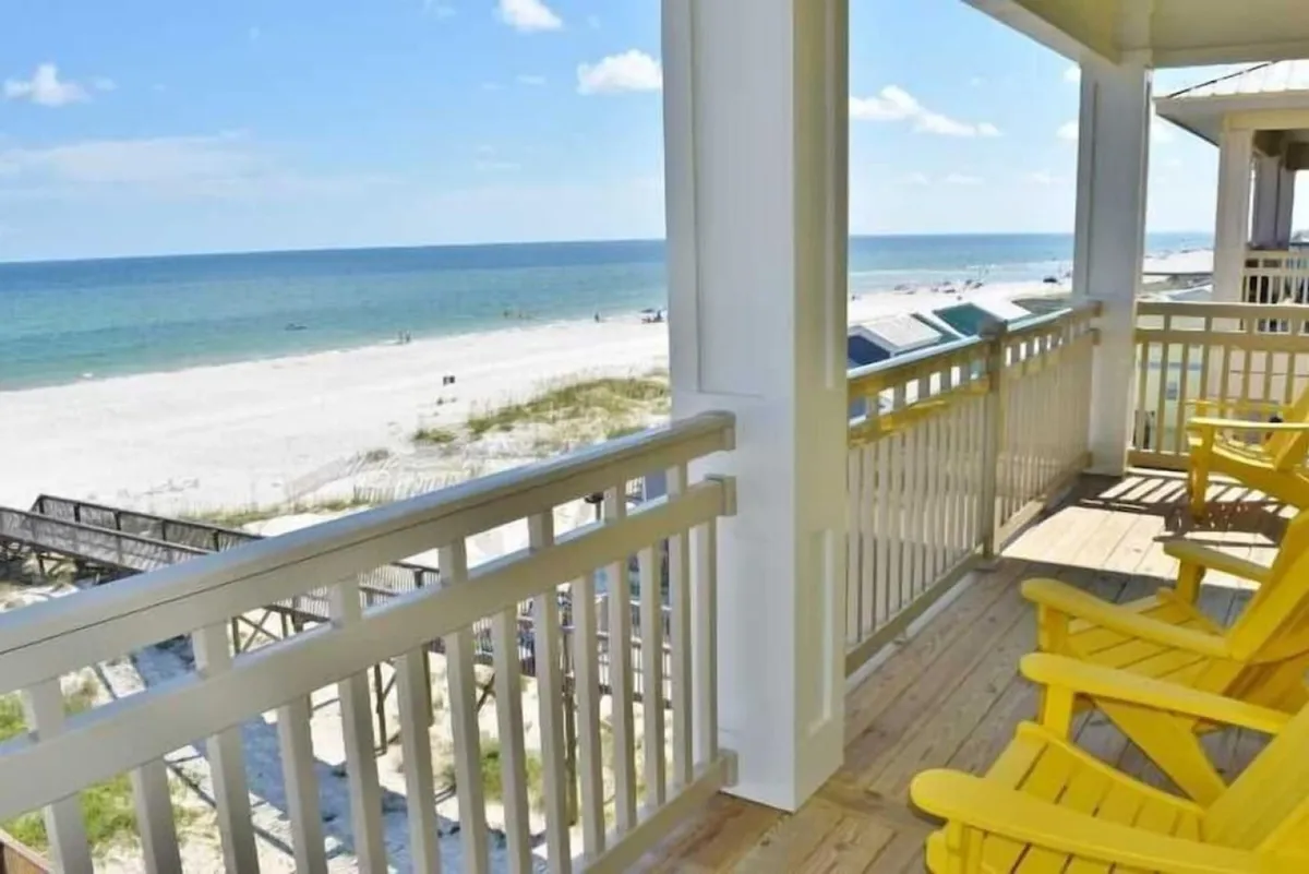 A beachfront porch with bright yellow Adirondack chairs overlooks sugar-white sands and the calm ocean. The wooden deck is bordered by a railing, and a dune walkover structure provides direct access to the beach, inviting guests to step right onto the sand.
