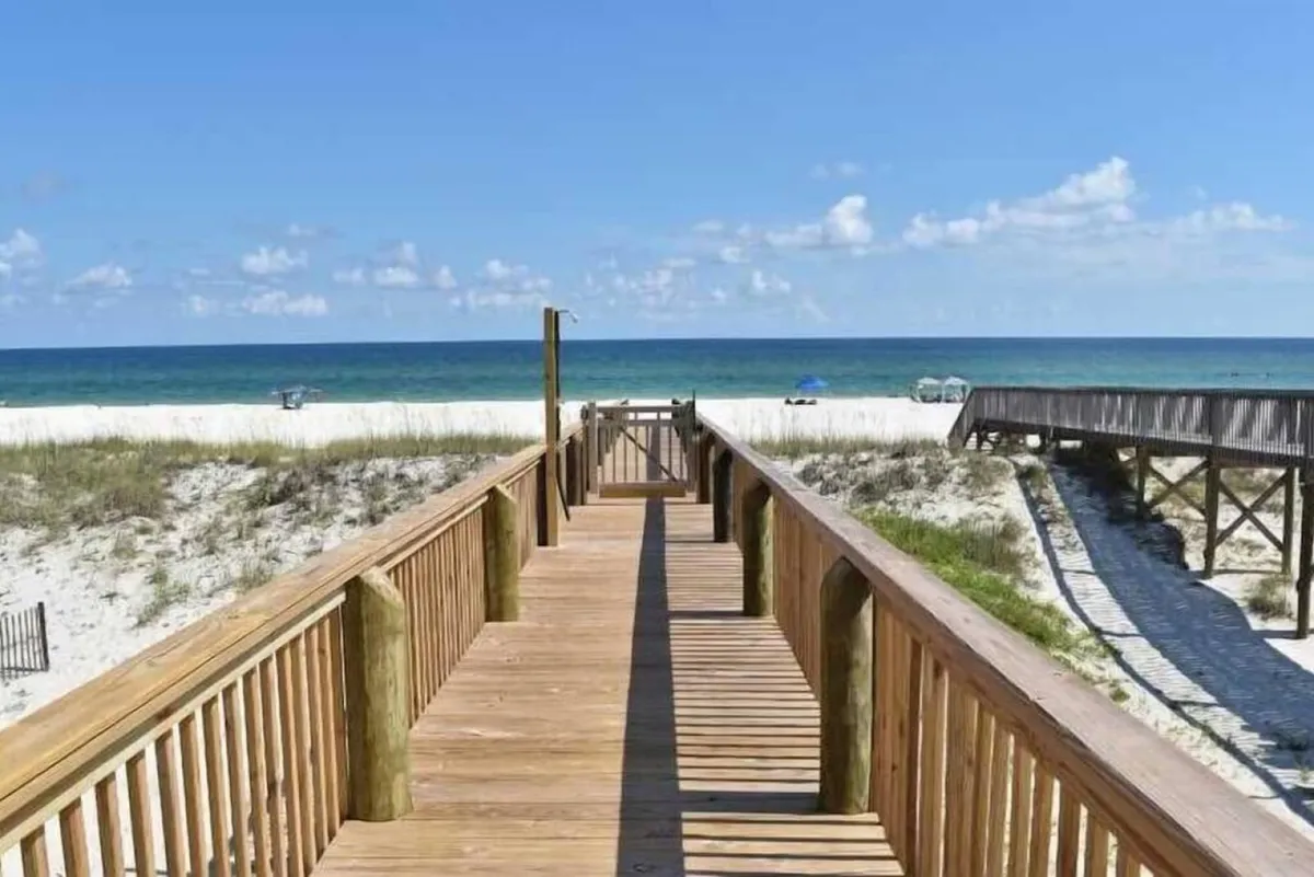 A wooden boardwalk stretches from the house directly to an expansive, pristine white sandy beach and emerald green ocean under a clear blue sky. The scene emphasizes unobstructed access to the shoreline and tranquil natural beauty, perfect for sunbathing, swimming, or sandcastle building.