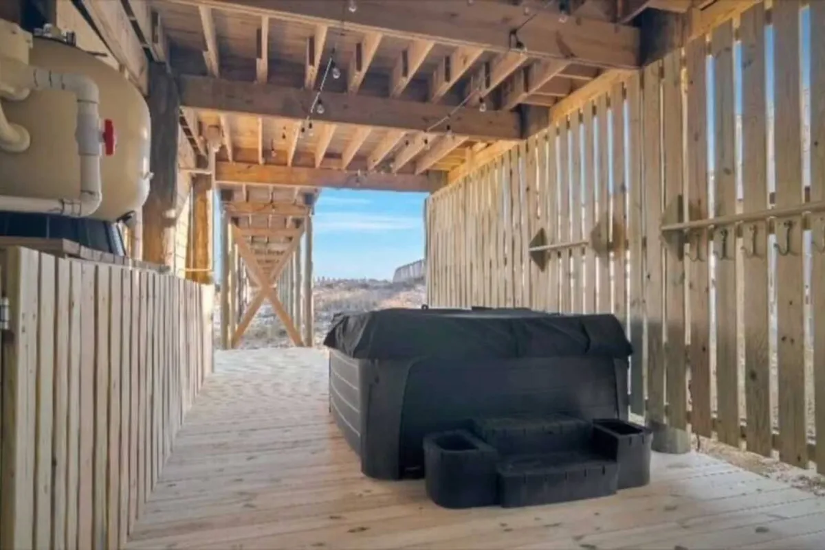 A secluded hot tub rests on a wooden deck, surrounded by a slatted privacy fence and shaded structural beams. The blue sky and sandy beach are visible just beyond the fence, emphasizing the tranquil, beachside setting. Hooks on the fence provide convenient spots for towels or swim gear, enhancing the sense of comfort and thoughtful design.