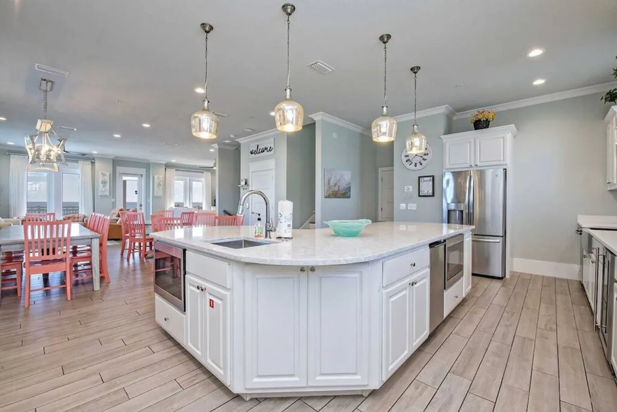 This image showcases a large white kitchen island with a built-in sink and appliances, surrounded by rows of coral-colored dining chairs and tables. Stainless steel refrigerators and hanging pendant lights frame the background, emphasizing the spacious open kitchen-dining layout. The setting is bright and airy, ideal for group meal prep, serving, and communal dining.