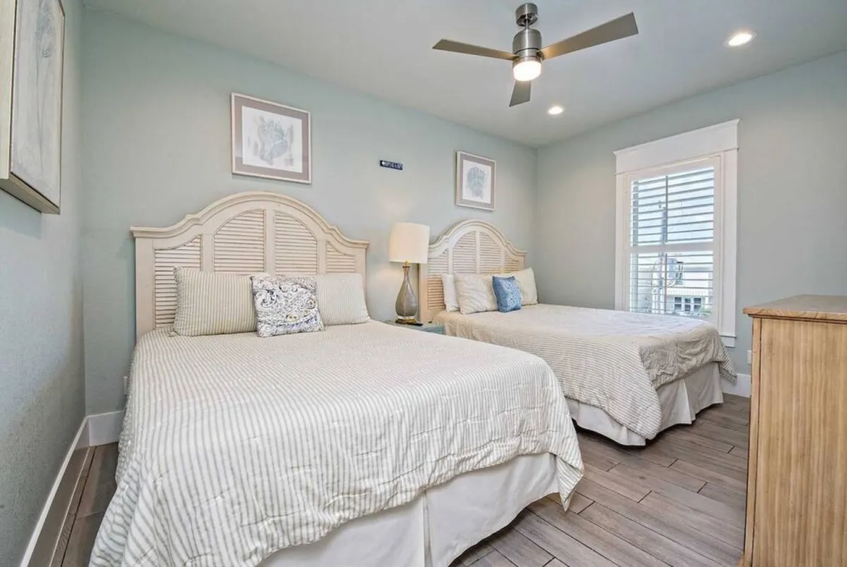 A spacious double-queen bedroom featuring two matching beds, a bedside table with lamp, ceiling fan, and a window with plantation shutters. The room exudes a peaceful, inviting atmosphere ideal for both adults and children, ensuring flexibility for group stays.