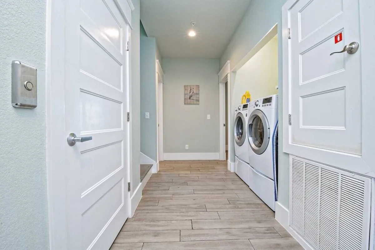 A laundry area with front-loading washer and dryer in a hallway lined with multiple doors, suggesting easy access from bedrooms. The scene is neat and functional, featuring wall art for a touch of hominess.