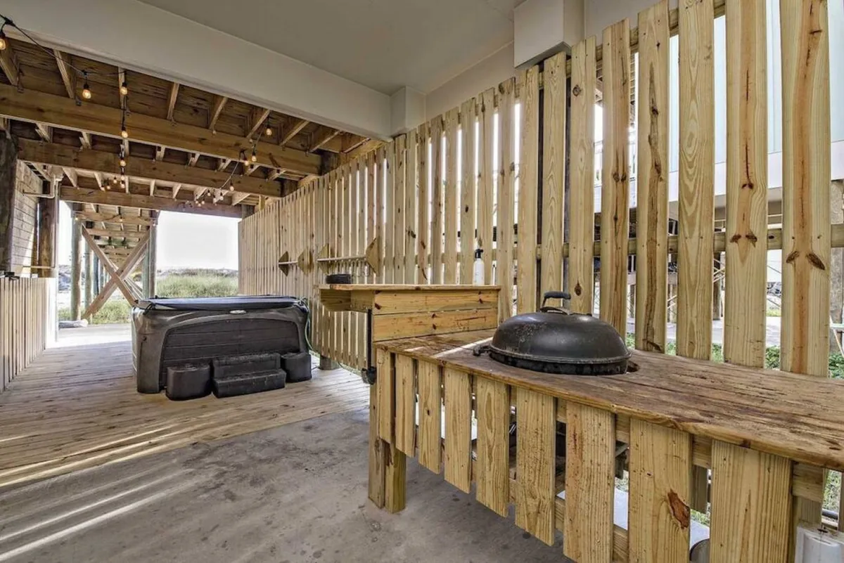 This cozy evening exterior scene features a hot tub, a wooden grill station, and rustic string lights overhead, all enclosed by a wooden privacy fence. The setup creates an intimate, inviting atmosphere ideal for casual gatherings, grilling, and enjoying evenings outdoors—potentially by the fire pit.