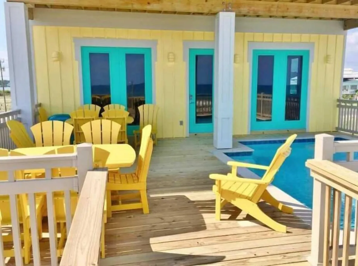 This vibrant exterior scene features bright yellow Adirondack chairs and a dining set on a deck, turquoise French doors opening onto the space, a private pool with sparkling blue water, and the yellow exterior wall of the house. It conveys an atmosphere of relaxed outdoor dining, poolside fun, and casual group gatherings with the beach just beyond.