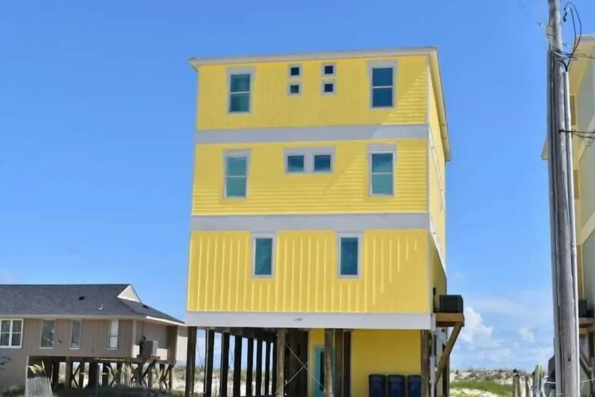 A tall, three-story yellow beach house on stilts stands out against the blue sky, with multiple windows and a backdrop of distant dunes. The scene suggests family vacations, outdoor gatherings on porches, and easy access to local shops and restaurants.