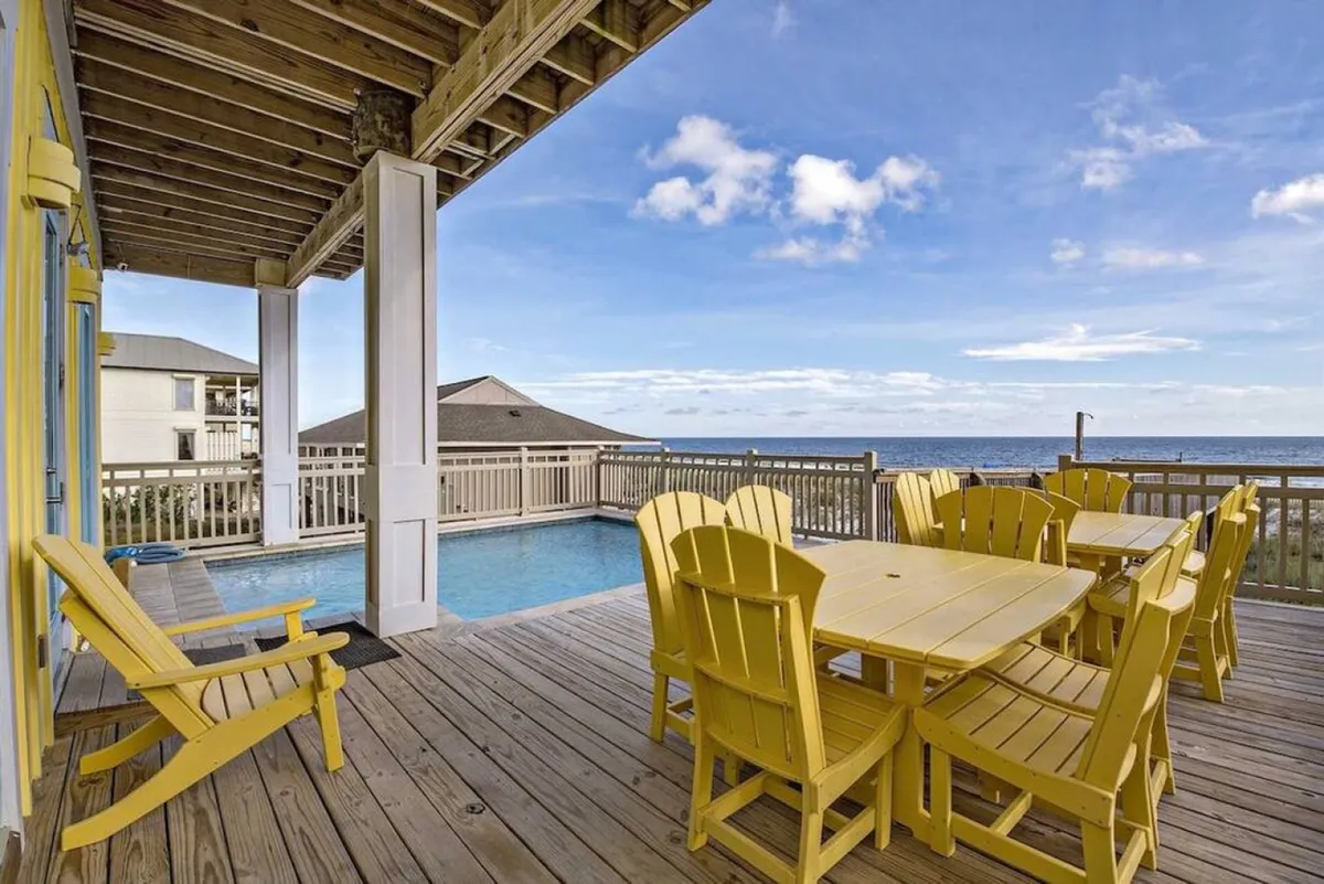 A bright exterior view captures the private heated pool, yellow Adirondack chairs and dining set, a wooden deck, and unobstructed ocean views. The beachfront railing hints at safety and exclusivity, while the setup invites both poolside sunbathing and outdoor dining with a backdrop of the sea.