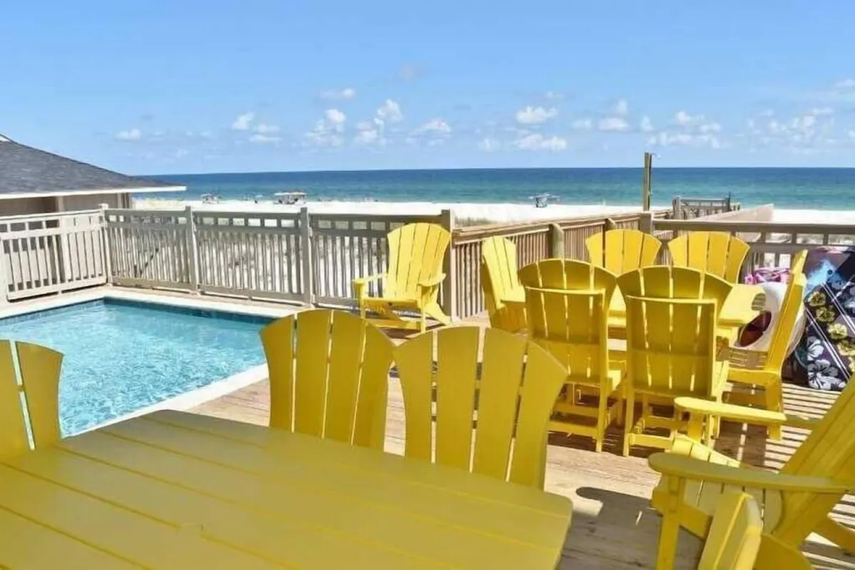 A cheerful outdoor scene with a private heated pool, yellow Adirondack chairs, a wooden deck, and a beautiful ocean view in the background. The space conveys a lively, social atmosphere ideal for gatherings and relaxation.