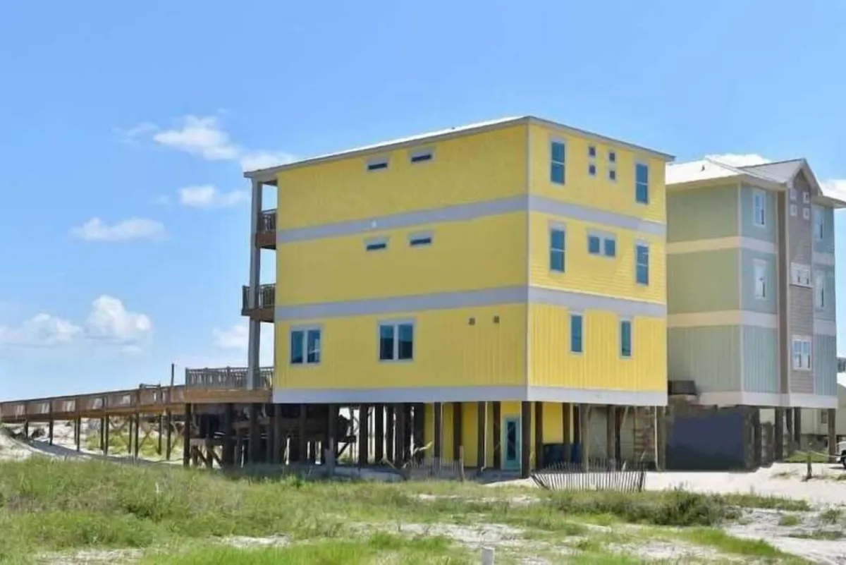 A wide exterior view of the bright yellow three-story beach house on stilts, with a wooden dune walkover leading toward the beach. The image also shows neighboring colorful houses, green dune grasses, and a spacious porch for gatherings or relaxation. The mood is vibrant and carefree, evoking the spirit of a private seaside retreat.