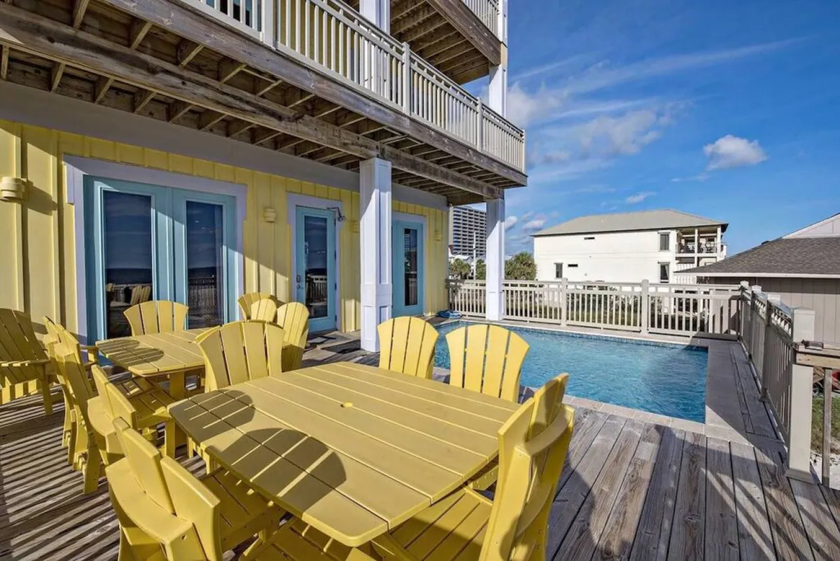 A bright and cheerful outdoor setting featuring a private pool with clear water, a yellow wooden dining table and chairs, and a spacious beachfront porch attached to the house. The backdrop is a sunny sky with scattered clouds, adding to the inviting, relaxed mood. The scene showcases spaces for swimming, sunbathing, outdoor dining, and gathering with family or friends.