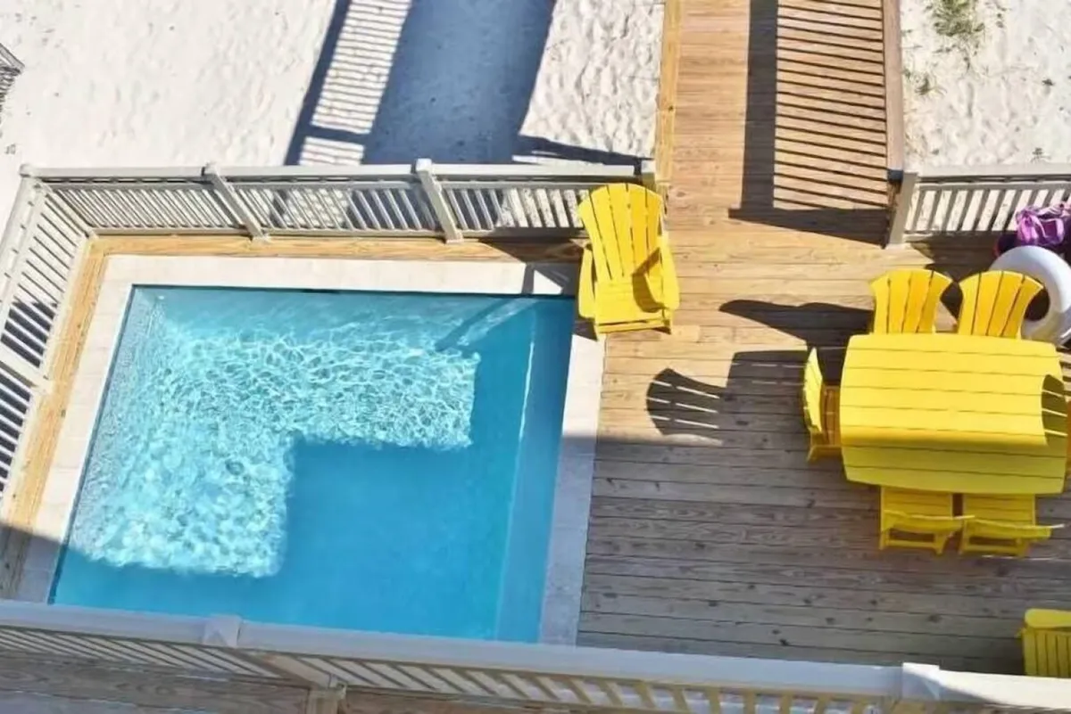 This vibrant exterior shot showcases the private heated pool with clear blue water, surrounded by bright yellow dining furniture on a wooden deck. The pool area is enclosed with white fencing and opens up to a sandy beach beyond, creating a seamless connection between poolside relaxation and beachfront access. The image also hints at a nearby fire pit area, inviting guests to imagine transitioning from daytime swimming to evening gatherings.